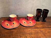 Group view of two red plates with matching cups and two black Treasure Island mugs