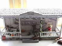Front view of Cucumber Cottage porch diorama showing structure, dolls, furniture, pots, pumpkins, bottle and garland