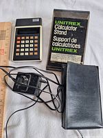 Calculator, adapter, stand in box, carry case laid out with ruler for scale