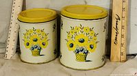 two tin canisters with lids on, rulers for scale