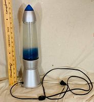 Full view of silver rocket-style lava lamp beside ruler showing ~16 in height with power cord and inline switch