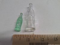 Clear and green miniature Coca-Cola bottles beside ruler showing scale