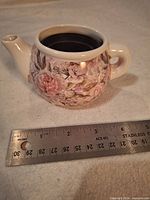 Overall view of ceramic teapot-shaped planter with floral design and ruler for scale