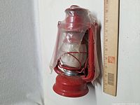 Red metal oil lantern beside ruler showing size, protective plastic wrap