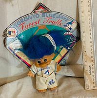 Front view of Blue Jays Forest Troll doll with blue hair, uniform and front of cardboard backing, ruler showing scale