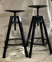 Front view of two black adjustable bar stools showing tripod bases and screw posts