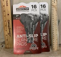 Front view of two sealed X-Protector 16-piece anti-slip furniture pad packs next to rulers for scale