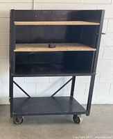 Front view of black metal rolling cart showing two plywood shelves and lower shelf on casters