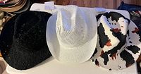 Three cowboy hats in black crochet, white crochet, and cow print fabric on table with tags visible