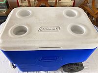 Blue wheeled cooler showing lid with Coleman logo and four cup holders
