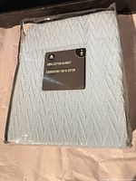 Front view of sealed package showing light blue cable knit blanket and brand insert card