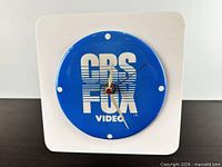 Front view of square clock showing blue CBS/FOX VIDEO dial and hands