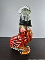 Left side view of clear-headed, amber/red glass rooster decanter with black raffia neck wrap