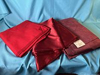 Three stacks of red fabrics—matte woven, shiny satin, suede-finish—set on blue cloth