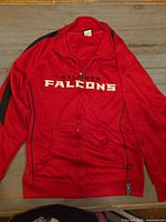 Front view of red Atlanta Falcons track jacket showing chest text, zipper and pockets