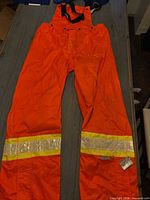 Full view of orange bib pants laid flat showing reflective bands and suspenders