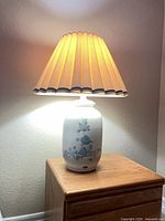 Full view of lamp illuminated, showing ceramic base, floral motif, pleated shade, wooden table for scale