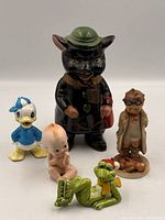 Group shot of five ceramic pieces: black pig bank, Hummel doctor boy, frog with skates, Kewpie, and Dewey Duck