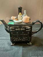 Side view of black stove-form teapot showing spout, handle, relief stove detailing and attached miniature cookware
