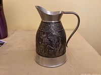 Side view of embossed pewter pitcher showing handle, spout, base and detailed relief artwork