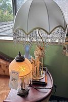 Both lamps on side table; figurine lamp with beaded dome shade behind smaller illuminated glass lamp