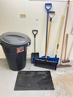 Overall view showing black garbage bin with lid, shovels, hoe and rubber mat