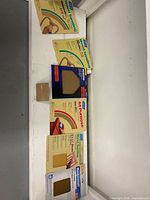 Overview of six unopened packs of Norton sandpaper sheets and one loose sandpaper block arranged on shelf