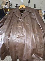 Front view of brown leather men's coat displayed on hanger showing three-button closure and pockets