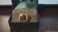 Front view of gold-tone ring with square dark gray stone in green case