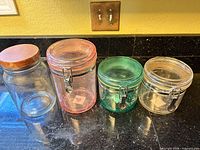Four glass storage jars on countertop: clear jar with wood lid, pink-tinted clamp jar, green-tinted clamp jar, clear clamp jar