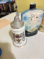 White ceramic München stein with pewter lid beside map-shaped decanter