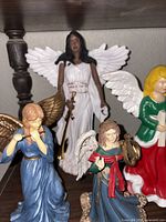 Shelf with four angel figurines including tall white-gowned angel and three smaller angels in blue and green-red outfits