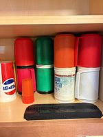 Pepsi cup, small orange cup, six colored insulated mug parts stacked showing four complete mugs, black rubber bar mat