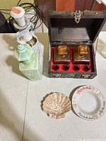 Lot overview showing Jim Beam decanter, celadon bottle, wooden box open with amber decanters and glasses, shell dish, Hamm's plate