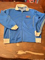 Full view of blue track jacket laid flat showing front, zipper, striped sleeves, contrast cuffs and hem