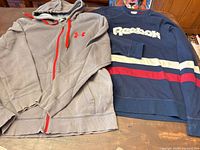 Under Armour hoodie and Reebok sweatshirt laid flat showing fronts