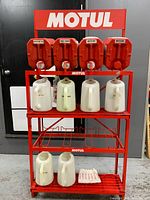 Front view of red Motul stand with 4 red 20 L jugs on top shelf, 3 white pouring jugs on middle shelf, 3 white pouring jugs on bottom shelf, white drip tray on lowest level