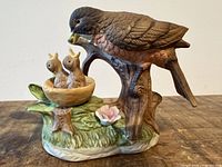 Front view of ceramic figurine showing mother bird feeding two chicks in nest on tree trunk base with leaves and flower