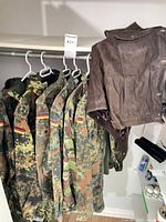 Multiple German Flecktarn garments and brown leather jacket on hangers