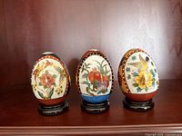 Front view of three cloisonné porcelain eggs on stands showing bird and floral motifs