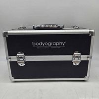 Front view of closed black Bodyography cosmetics train case showing branding, latches and handle