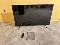 Front view of Samsung 43" QLED TV on stand with remote and manual on floor