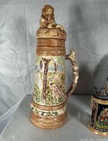Tall hunter stein with lion finial, raised relief deer scene left view