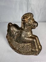 Front right view of gold-tone rocking horse coin bank showing coin slot and floral base