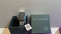 Watch in open box alongside lid showing Nic & Syd logo