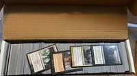 Open cardboard long box packed with Magic: The Gathering cards, three sample cards laid on top