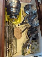 Drawer showing nested stainless pots/bowls, cooling rack, woven trivet, pans, colander, tongs, oven mitts