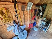 Overall view of shed wall showing shovels, trimmer, extension cord, dustpan, trash can, shelving with containers
