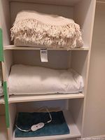 Shelf with cream fringed throw, white bedding piece, blue heating pad with controller visible