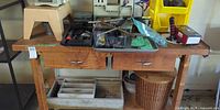 Bench top showing assorted hand tools, grey tray, yellow bin, stool and wicker basket under bench